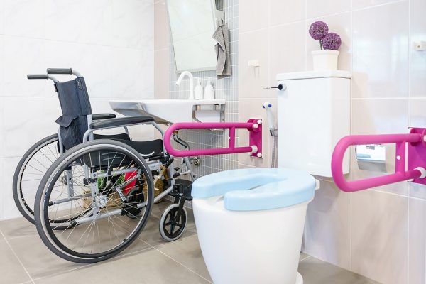 An accessible bathroom in someone's home. It shows a handrail on either side of the toilet and a raised hand-basin allowing a wheelchair to pull up to use it.