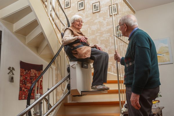 An older woman sits on a chair lift going up stairs in her house, with her husband watching.