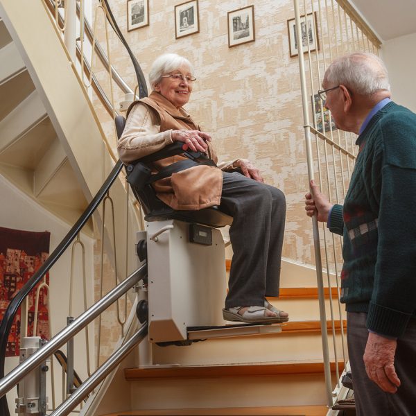 An older woman sits on a chair lift going up stairs in her house, with her husband watching.